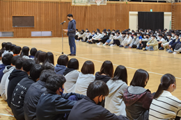 岡谷南高校の大体育館で行われた対面式にて、マイクの前に立つ藤澤校長先生の話を、体育館いっぱいに座った全校生徒が真剣に聞いている様子。