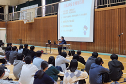 岡谷南高校の体育館で行われた交通安全講話の様子。岡谷警察署の竹内交通課長を講師に迎え、自転車の交通ルールやヘルメット着用の努力義務化について、スクリーンを使いながら1年生に説明している場面。
