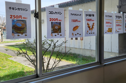 岡谷南高校の窓に掲示された昼食販売のメニュー表。チキンカツパンやコロッケパンなど、生徒に人気のパンメニューが価格付きで紹介されている。
