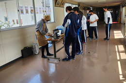 岡谷南高校の廊下にて、マンモウ食堂の販売スタッフからお弁当やパンを購入したり、次回の予約をしたりする生徒たちの賑やかな風景。