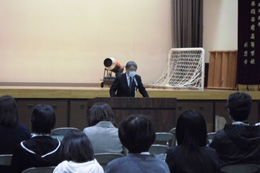 岡谷南高校の小体育館で開催されたPTA総会にて、演台で挨拶をする役員と、その話を真剣に聞く保護者たちの後ろ姿。