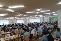 岡谷南高校の生徒たちが研修先の広い室内で、クラスごとに整列して昼食や休憩をとっている様子。