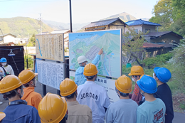 岡谷南高校の生徒たちがヘルメットを着用し、松代大本営・象山地下壕の入り口付近で案内板を見ながらガイドの説明を受けている場面。