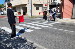 岡谷南高校の交通安全街頭指導にて、横断歩道で赤い旗を持ち、自転車で通学する生徒の安全を見守る職員の様子。
