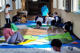 岡谷南高校の廊下にて、ブルーシートを敷いて大きな模造紙に背景画を描き上げる生徒たちの様子。