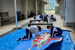 岡谷南高校の屋外スペースで、床に広げた大きな布や板にペイントを施し、文化祭の巨大装飾を作る生徒たち。