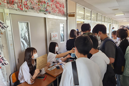 岡谷南高校の南高祭にて、クラス展示の受付で来場者を笑顔で迎える生徒たちと、賑わう廊下の風景。