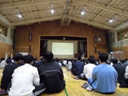 岡谷南高校の体育館にて、前方のスクリーンを見上げながら教育実習生による進路の話を聴く2年生の様子