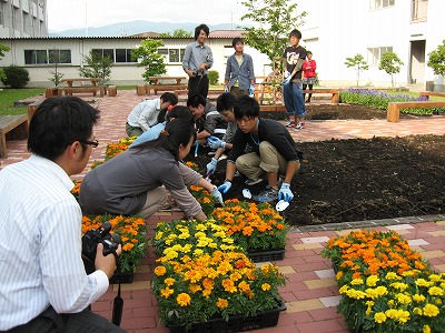 岡谷南高校：カメラを向ける教職員の前で、腰を据えて一つひとつ丁寧に苗を土に植えていく生徒たち。