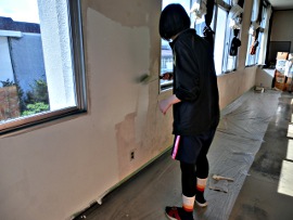 岡谷南高校：教室の壁の塗装作業。ローラーを使って、窓際から順に新しい塗料を慎重に塗り広げている生徒の後ろ姿。