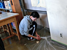 岡谷南高校：塗装の下準備として、床や塗装しない壁の境界にビニールとマスキングテープを使い、丁寧に養生作業を行っている生徒の様子。