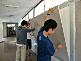 岡谷南高校：被服室前の廊下にて、整美委員の生徒たちが協力して大きな掲示板の素材板を壁に掲げ、位置を合わせながら仮止めをしている様子。