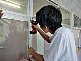 岡谷南高校：掲示板の設置作業。電動ドライバーを使い、掲示板の四方を固定するための金具やネジを慎重に取り付けている生徒の横顔。