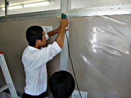 岡谷南高校：掲示板設置の仕上げ作業。壁に電動ドリルで穴を開け、作品展示に耐えられるよう頑丈に掲示板を固定している様子。