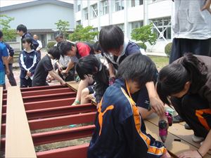 岡谷南高校：中庭ステージ設置作業。生徒会役員や有志の生徒たちが、鉄骨の土台に床材を並べ、電動工具などを使って一枚ずつ丁寧に張り付けている様子。