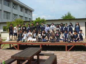 岡谷南高校：中庭ステージ完成記念の集合写真。作業に参加した生徒、職員、PTA、業者の皆さんが、新しく完成した木製ステージの上に並んで笑顔で収まっている様子。