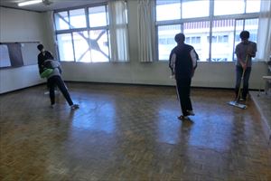 岡谷南高校：中性洗剤で拭き上げた後の教室で、複数の生徒たちが協力しながら床全体にワックスを均一に塗り広げている様子。