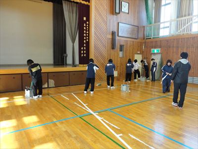 岡谷南高校：小体育館にて、女子バスケットボール部の生徒たちがステージ前から入り口に向けて、専用モップでワックスを塗布している様子。