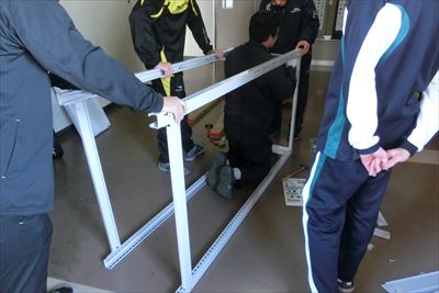 岡谷南高校：整美倉庫にて、生徒たちがスチール製の大きな棚のフレームを垂直に立て、組み立てを開始している様子。