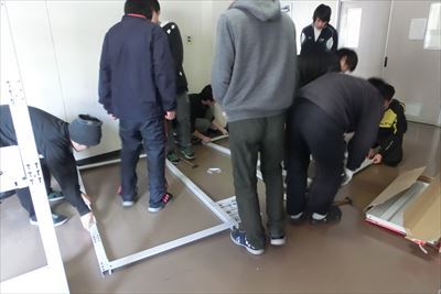 岡谷南高校：倉庫の床に部品を広げ、複数人の生徒たちが協力して棚の土台部分を慎重に組み上げている様子。
