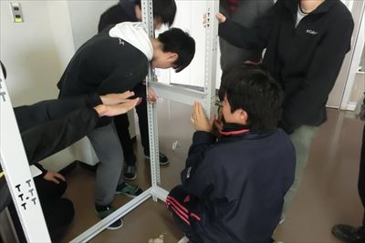 岡谷南高校：棚の水平を確認しながら、ネジを締めて固定作業を行う生徒たちの真剣な表情。