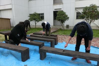 岡谷南高校：中庭整備にて、青いシートの上で生徒たちが刷毛を使い、木製ベンチに保護塗料を丁寧に塗っている様子。