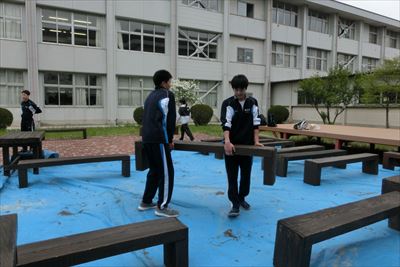 岡谷南高校：中庭整備にて、男子生徒たちが二人一組で協力し、塗装を終えた重い木製ベンチを運搬・設置している様子。