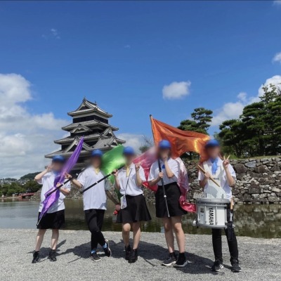 岡谷南高校の生徒たちが、セイジ・オザワ 松本フェスティバル（OMF）歓迎パレードの際、松本城を背景にカラフルなフラッグや楽器を持って記念撮影をしている様子。