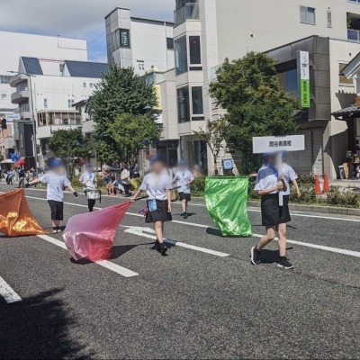 岡谷南高校のプラカードを掲げ、松本市内の道路を鮮やかなフラッグを振りながら行進するパレードの風景。