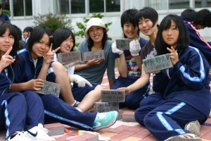 岡谷南高校：インターロッキングブロックの設置。裏面に思い思いのメッセージを書き込んだブロックを手に、作業中の女子生徒たちが笑顔で見せ合っている記念写真。