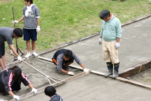 岡谷南高校：中庭整備の基礎作業。専門業者の指導のもと、生徒たちがトンボや木材を使って、砂を平らにならす「砂ならし」を行っている様子。
