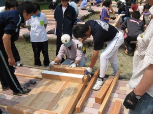 岡谷南高校：テーブルの組み立て作業。保護者や学校関係者に見守られながら、生徒が電動ドライバーを使って木材を固定している様子。