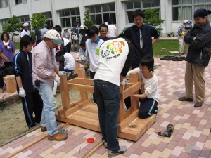 岡谷南高校：グループに分かれてのベンチ製作。設計図を確認しながら、大きな木材を組み合わせて土台を作る生徒と大人の協力風景。