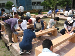 岡谷南高校：中庭に並べられた多数のベンチ。多くの生徒や保護者が参加し、一斉にやすりがけや防腐処理を行っている活気ある光景。