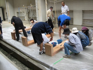 岡谷南高校：仕上げのニス塗り作業。地面に汚れがつかないようビニールを敷き、完成したベンチに丁寧に刷毛でニスを塗っている様子。